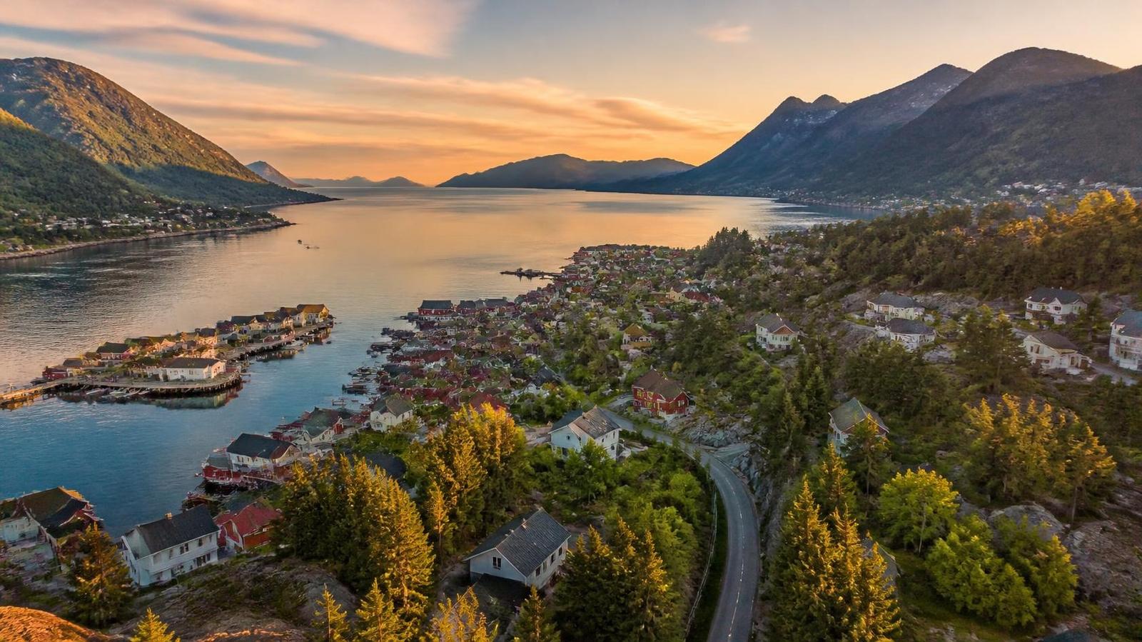 Nordiskt kustlandskap med fjord, skog och en liten hamnstad i kvällsljus